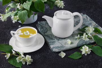 Still life with green tea in a white cup, white teapot on a dark background. Fresh jasmine flowers in a vase as a decoration
