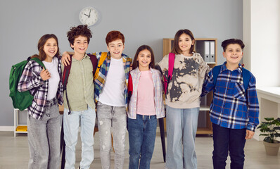 Group of happy junior school students standing in classroom. Cheerful schoolchildren, friends in casual clothes and backpacks posing for group portrait at school. Smiling cool kids boys and girls