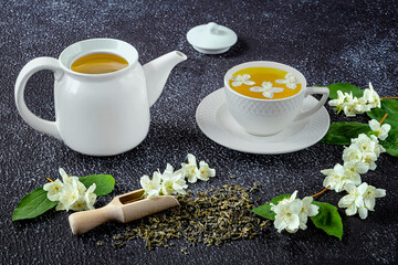 Green tea with jasmine in a white cup and teapot on a dark background. Fresh jasmine flowers and a wooden spoon as decor