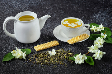 Green tea with jasmine in a white cup and teapot on a dark background. Fresh jasmine flowers and waffle dessert as decoration