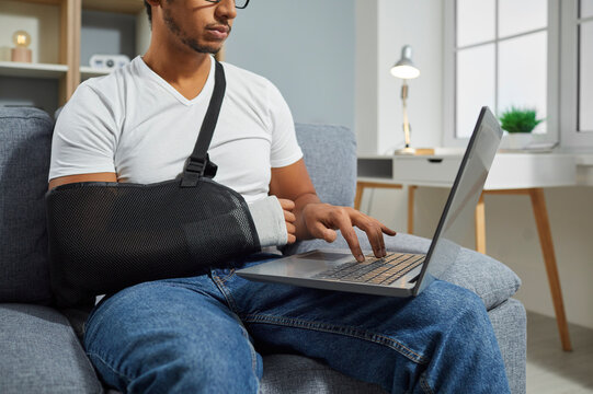 Cropped Shot Of A Young Man Wearing An Orthopedic Arm Sling On His Broken Wrist Sitting On The Couch At Home, Working On His Modern Laptop Computer, And Typing With His Left Hand On The Keyboard