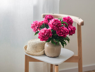 The interior of the living room is a bouquet of red peonies in a ceramic vase and a straw hat on a wooden chair in Scandinavian style