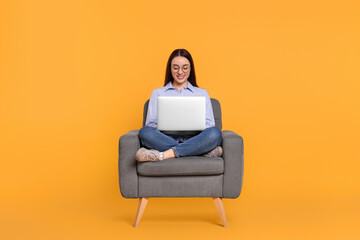 Smiling young woman working with laptop in armchair on yellow background