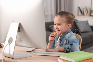 Little girl eating biscuit while using computer at table in room. Internet addiction