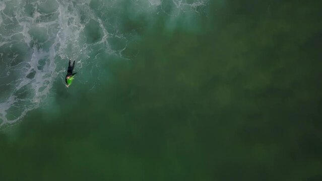 Young male surfer rowing on surfboard in crystal ocean water at scenic tropical beach on sunny day aerial view. Surfing man enjoying sport paddling and surfing on summer vacation drone shot.