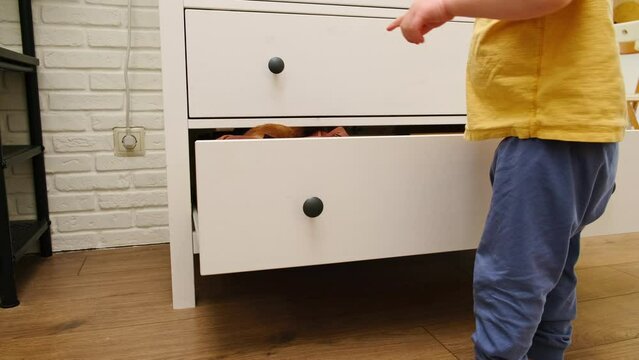 The toddler baby reaches into an open drawer of the chest. A child playing indoors opens a cabinet. Kid aged about two years (one year eleven months)