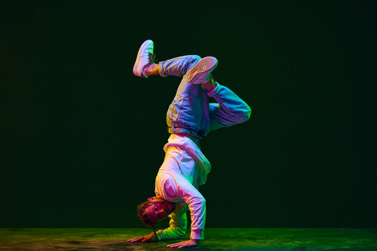 Young artistic man in sportive clothes dancing breakdance, hip-hop, street dance against black studio background in neon light