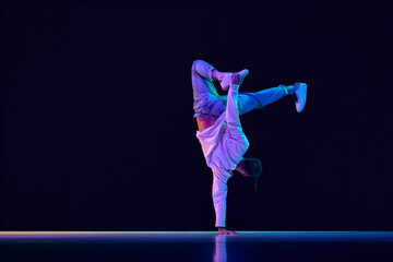 Youth culture. Young man in casual clothes making performance, dancing breakdance against black studio background in neon light