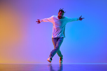 Young handsome guy in casual clothes and dreads dancing breakdance against gradient multicolored studio background in neon light