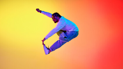 Dynamic image of young guy dancing breakdance against gradient orange yellow studio background in neon light