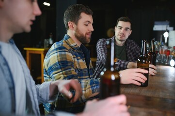 Three young men in casual clothes are smiling, holding bottles of beer while standing near bar counter in pub