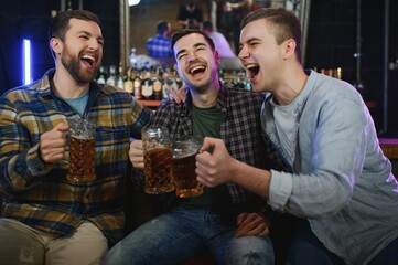 Three young men in casual clothes are smiling, holding bottles of beer while standing near bar counter in pub