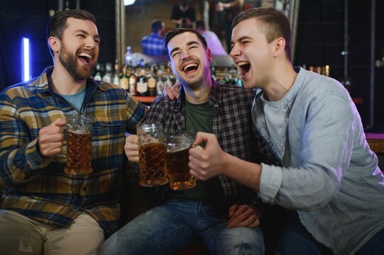 Three young men in casual clothes are smiling, holding bottles of beer while standing near bar counter in pub