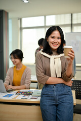 An attractive Asian female graphic designer stands in the office with a coffee cup in her hand