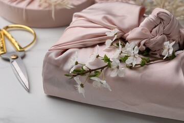 Furoshiki technique. Gift packed in pink fabric, beautiful flowers and scissors on white table, closeup