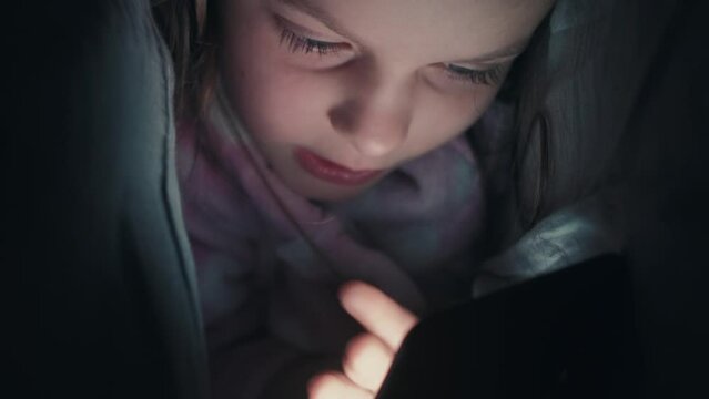 Vertical video. Bedtime phone. Sleepless child. Night online. Curious small girl using gadget under blanket surfing Internet late in bed in dark.