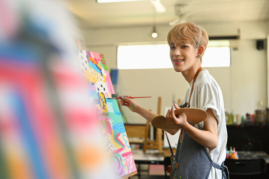 Happy asian student man in apron holding palette and painting on easel at art class. Education, hobby, art concept