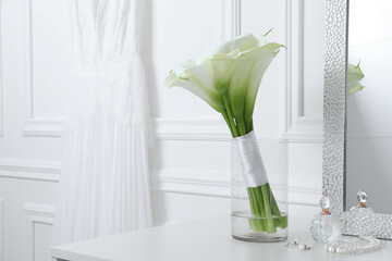 Beautiful calla lily flowers tied with ribbon in glass vase, bottle of perfume and jewelry on white table. Space for text