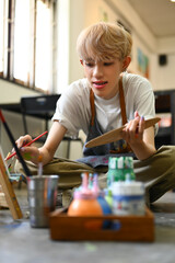 Image of young male artist holding palette and painting picture on canvas with oil paints in art studio