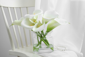 Beautiful calla lily flowers in glass vase on white chair indoors