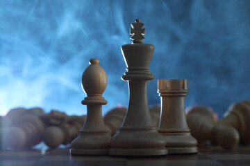 Wooden king, rook and bishop in front of fallen chess pieces on checkerboard against blue background, closeup