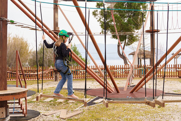 Teenage teen girl in climbing harness equipment, green sports safety helmet. Rope amusement park. Fastening attaching carabiner to safety rope. Hanging obstacle course. Kids children's attraction