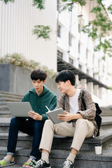 Fototapeta premium Two smart young Asian college students focusing on his school project, looking at laptop and tablet, discussing and working together at the campus park .