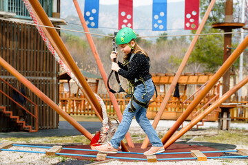 Teenage teen girl in climbing harness equipment, green sports safety helmet. Rope amusement park....