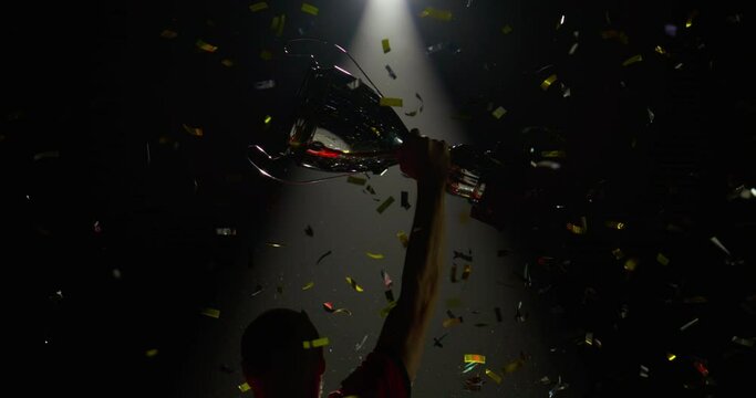 Silhouette of Caucasian male soccer football player raising a trophy above head against bright light and falling confetti. Super slow motion, shot on RED cinema camera
