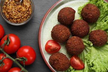 Delicious falafel balls, tomatoes, lettuce and grain mustard on dark grey textured table, flat lay. Vegan meat products