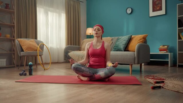 A young attractive woman sits in a lotus position, with her eyes closed, takes a slow breath in and out. Portrait of redhead woman practicing yoga at home. Sports concept. Yoga class. Slow motion.