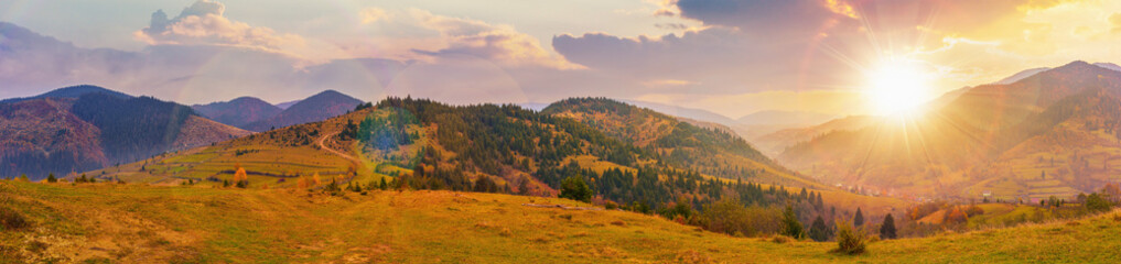 panorama of countryside landscape at sunset. grassy pasture meadows and forested hills in autumn. mountain ridge in the distance. village in the valley. clouds on the sky in evening light