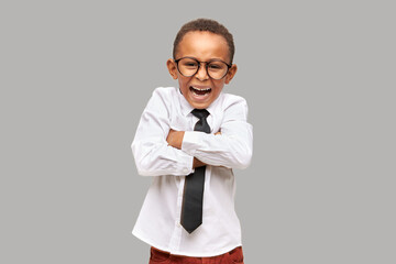 Little african american boy standing against gray studio background with folded arms shouting with frustration, upset with results of exams, dressed in white shirt and black necktie