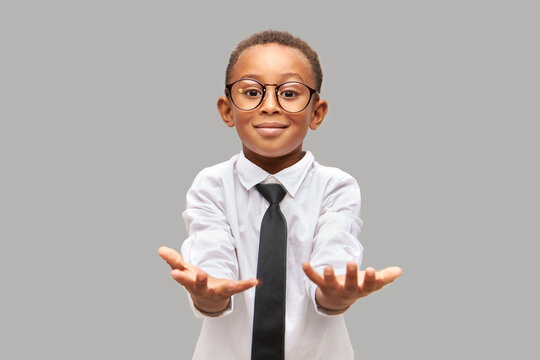 Studio portrait of cute happy enthusiastic excited schoolboy reaching opened hands to camera like holding something, ready to take knowledge, wearing eye glasses and school uniform with necktie