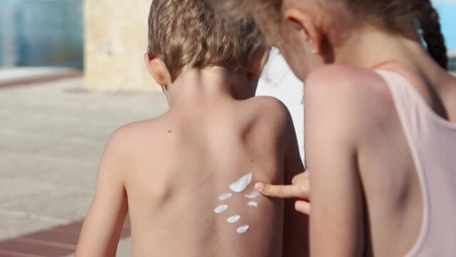 A girl puts sunscreen on her little brother's back.The girl draws the sun on the boy's back with suntan lotion.Protecting children on a sunny day.Family vacation on the beach.Cute brother and sister