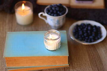 Cup of blueberries, set of books, phone, eyeglasses, lit candles and blanket on the table. Hygge at home. Selective focus.