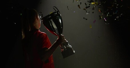 Silhouette of Caucasian woman female soccer football player raising a trophy above head against bright light and falling confetti. Super slow motion, shot on RED cinema camera