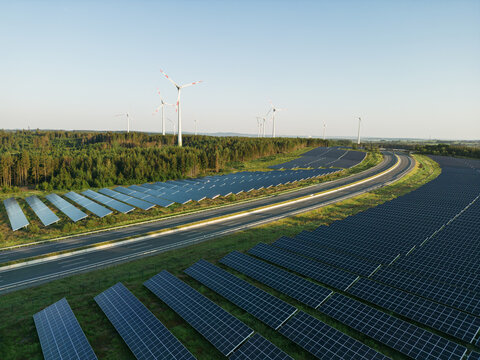 Autobahn zwischen Photovoltaik-Anlagen mit Windrädern im Hintergrund in grüner Natur bei Sonnenschein