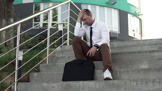 An upset man trying to calm down by breathing into a paper bag. Businessman suffering from panic attacks on the street, sitting on the city stairs.