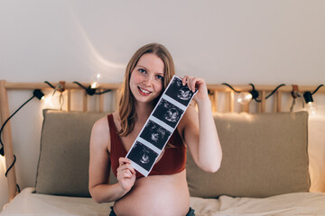portrait of happy caucasian pregnant woman sitting on bed showing ultrasound