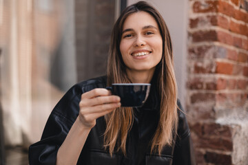 Close-up portrait of laughing blonde girl in black shirt with coffee in coffee shop terrace. Lovable lady sitting near window and holding cup of coffee and look happy. Woman drink, hold coffee mug.