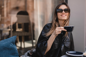 Close-up of laughing blonde girl in black shirt, glasses with coffee in coffee shop terrace. Lovable lady sitting near window and holding cup of coffee and look happy. Woman drink, hold coffee mug.