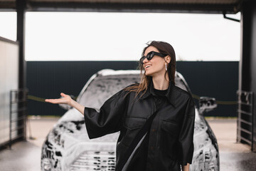 Stylish woman at car wash standing near transport car in foam soap. Soaping auto by professional...