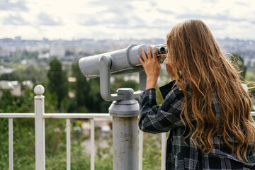 Back shot of a blonde woman looking to the city through some big binoculars.