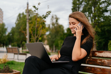 Cheerful woman is sitting on a bench talking on the phone and typing on laptop.