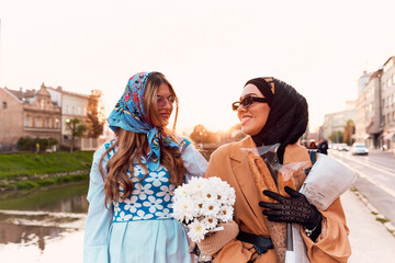 Couple woman one wearing a hijab and a modern yet traditional dress, and the other in a blue dress and scarf, walking together through the city at sunset. One carries a bouquet and bread, while the