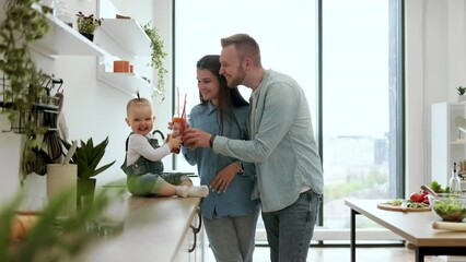 Adorable family dressed in denim attire toasting fresh drinks while resting in white spacious kitchen. Attentive parents taking care of little daughter while staying at home. Concept of love.