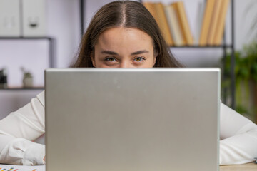 Confident business woman hiding behind laptop computer, looking at camera, spying his colleagues...