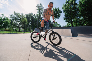 A mature shirtless tattooed man is exercising freestyle bmx tricks in a skate park.