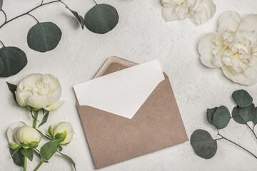 Fototapeta na wymiar Invitation or greeting card mockup with craft envelope, white peony flowers and eucalyptus sprigs on light background.